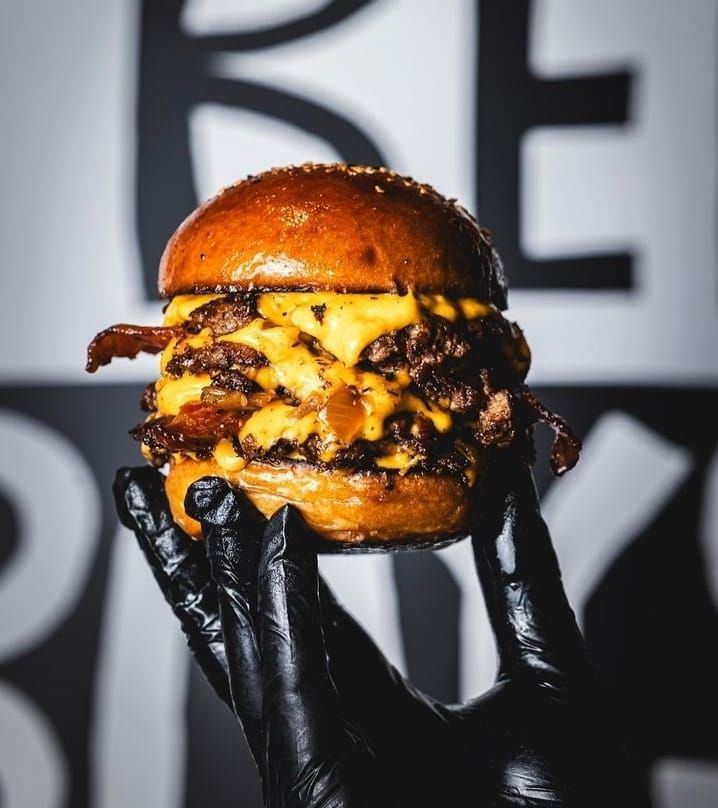 Mão com luva preta segurando um burger duplo com muito cheddar e bacon