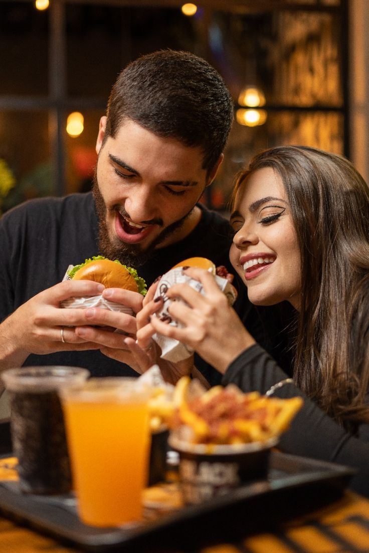Casal sorrindo enquanto come hambúrgueres, com batata frita com bacon e bebidas na bandeja