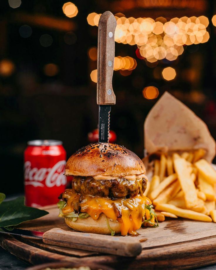 Burger com cheddar escorrendo e faca espetada no pão, com batata frita e refrigerante ao lado