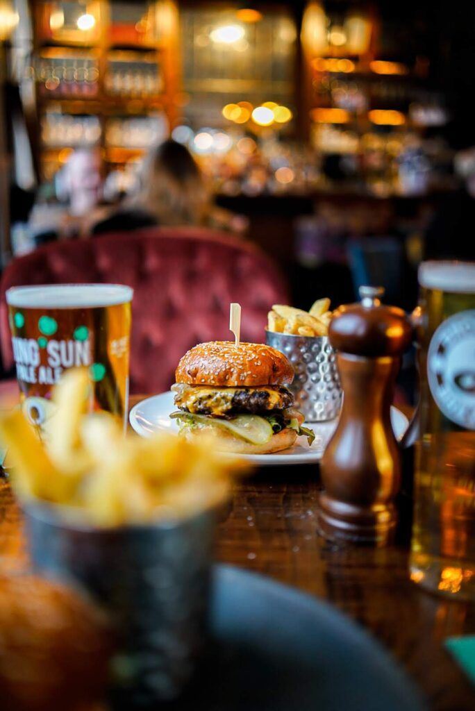 Burger com cheddar e picles servido na mesa do salão, com chope e batata frita ao fundo