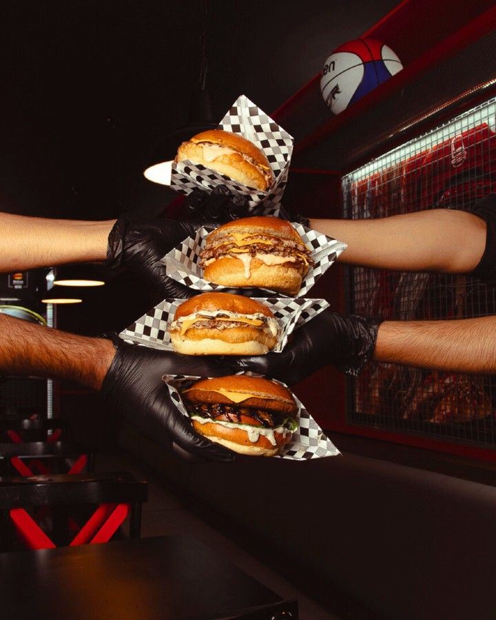 Quatro burgers em papel xadrez segurados por quatro mãos de luva preta, uma acima da outra