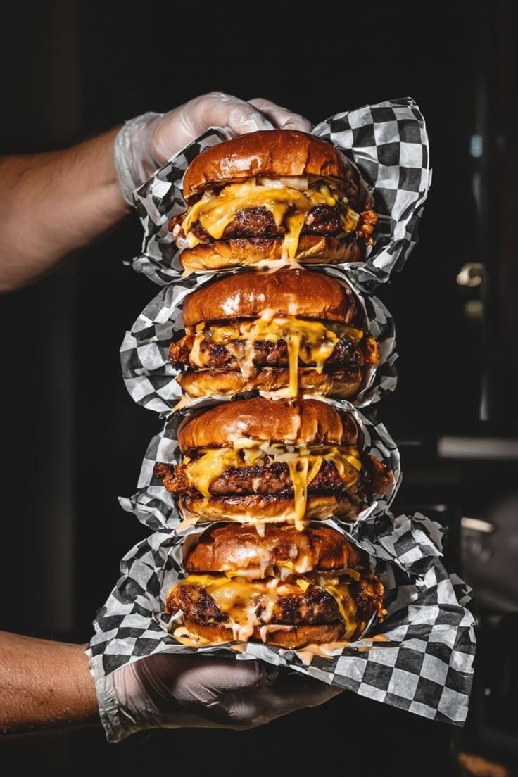 Quatro smash burgers empilhados com cheddar derretido, embrulhados em papel xadrez preto e branco, segurados por mãos de luva